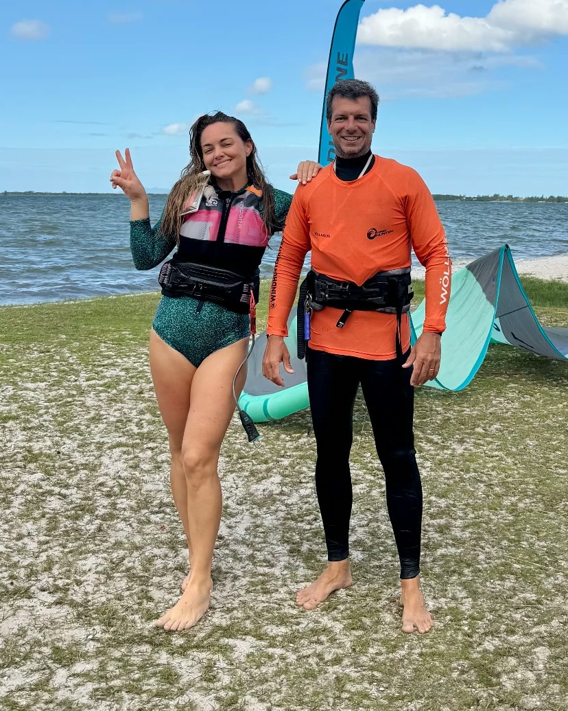Manu e um instrutor sorrindo na beira da lagoa, com colete e trapézio de kite